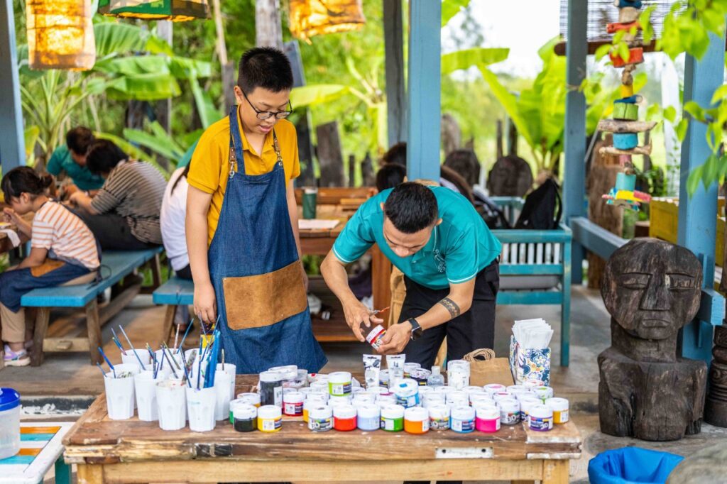 Participants engaged in a wood carving and painting workshop at Lang Cui Lu - Driftwood Village in Hoi An, Vietnam, learning traditional techniques from local artisans. The workshop showcases intricate driftwood art pieces, highlighting the craftsmanship and creativity of the artisans.